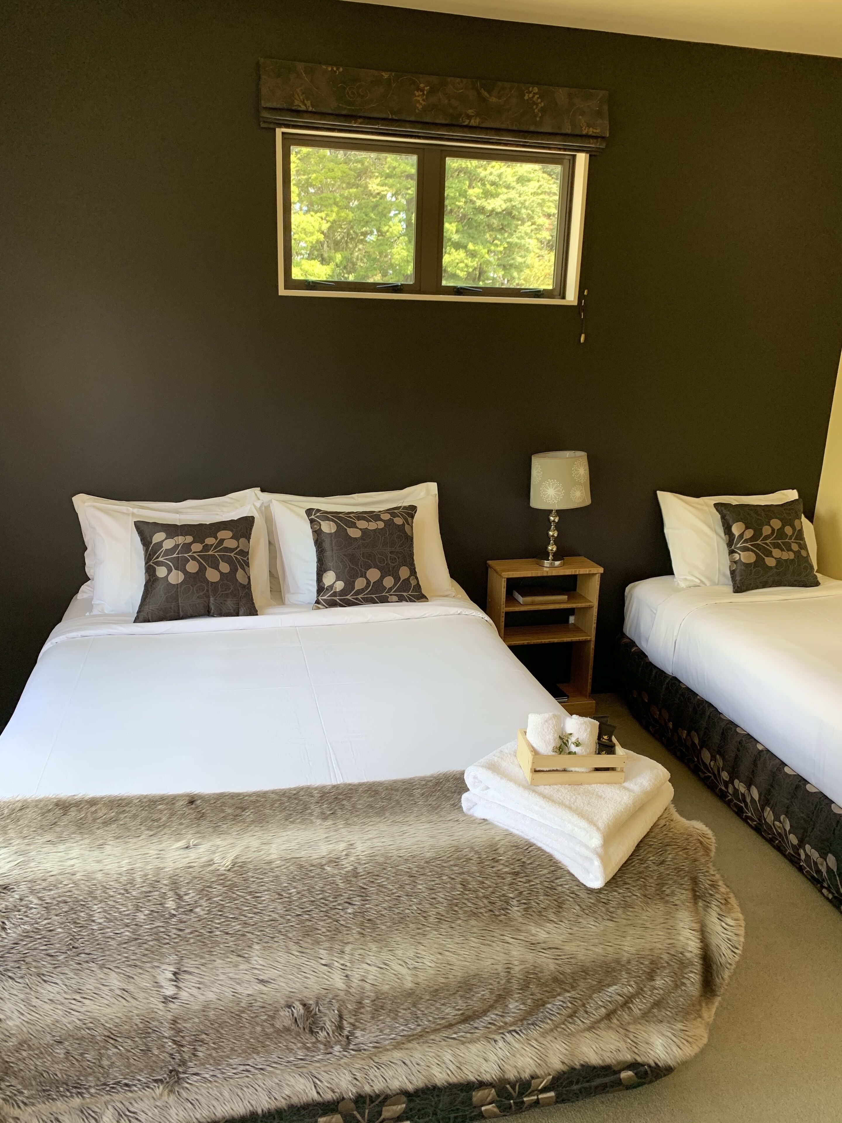 A hotel room with two beds, a nightstand, a lamp, and a window with a view of trees.
