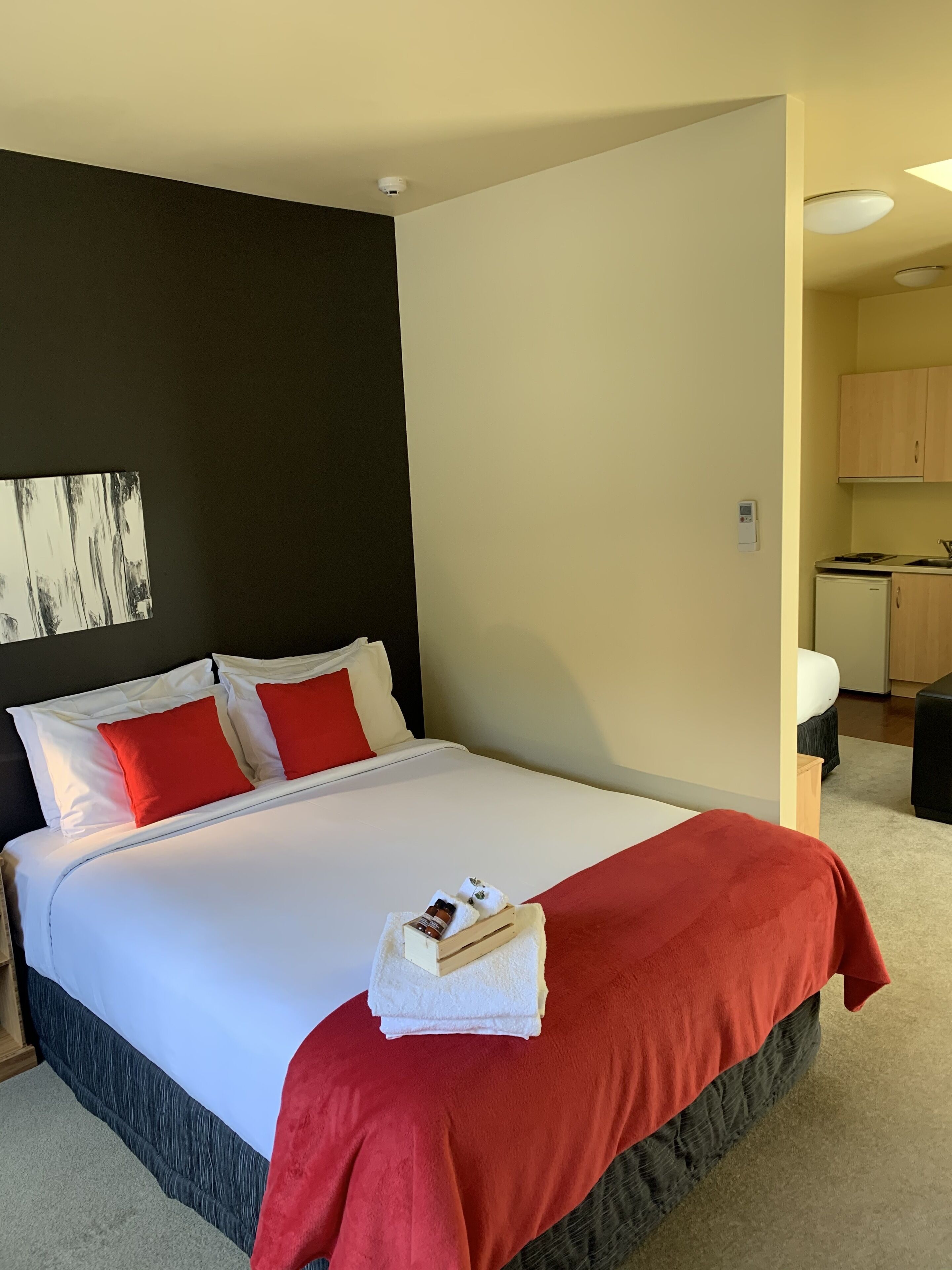 A hotel room with a large bed, a red and white bedspread, a small table with a bottle and a book, a white wall with a smoke detector, and a kitchenette area.