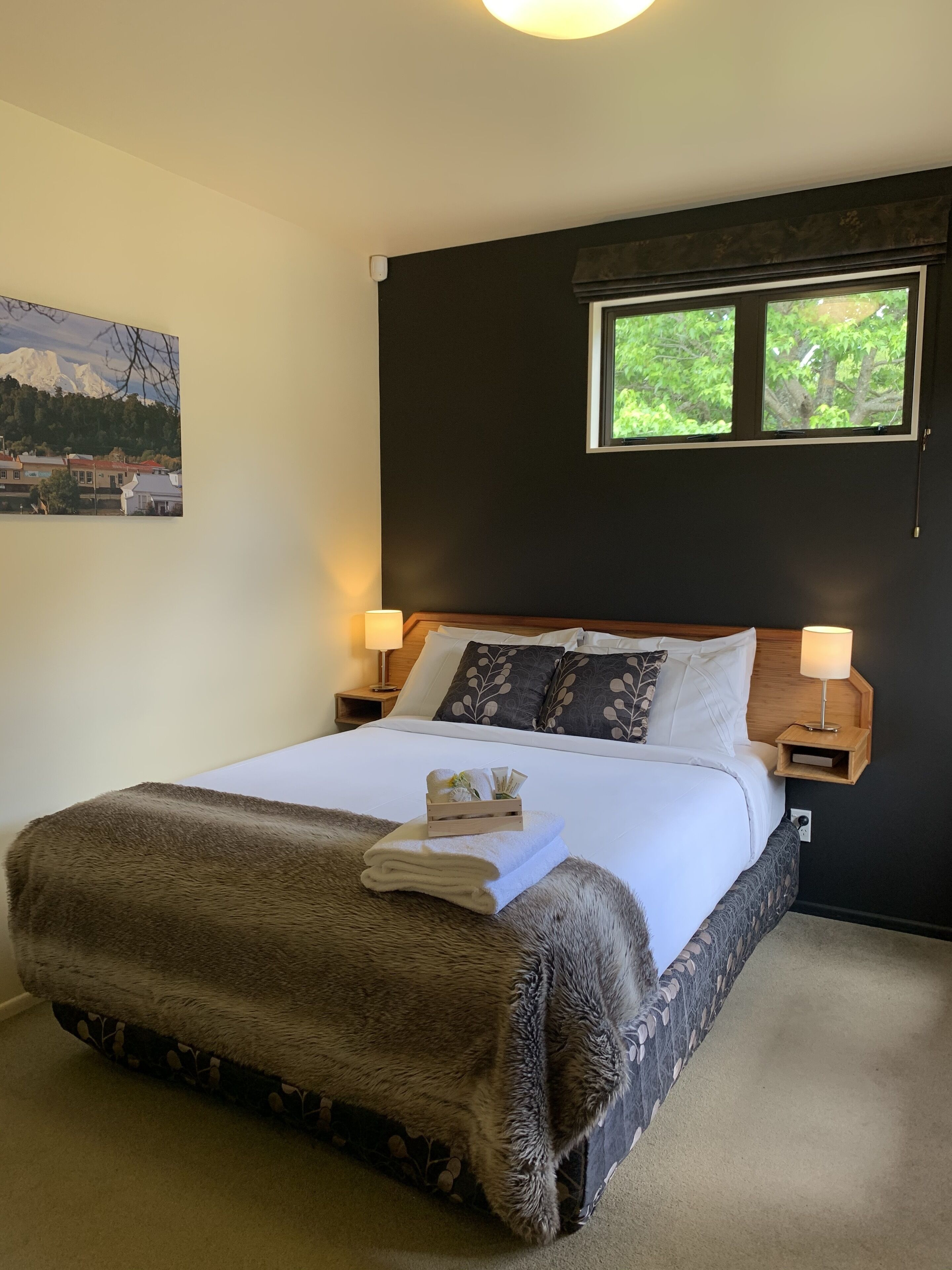 Una cama bien tendida con un edredón mullido y una pequeña bandeja de madera en el pie cama. Hay dos lámparas de noche y una ventana sobre la cama.