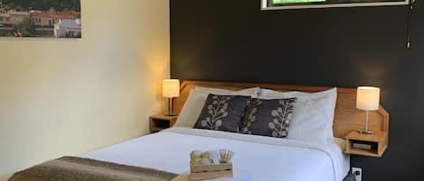 Una cama bien tendida con un edredón mullido y una pequeña bandeja de madera en el pie cama. Hay dos lámparas de noche y una ventana sobre la cama.