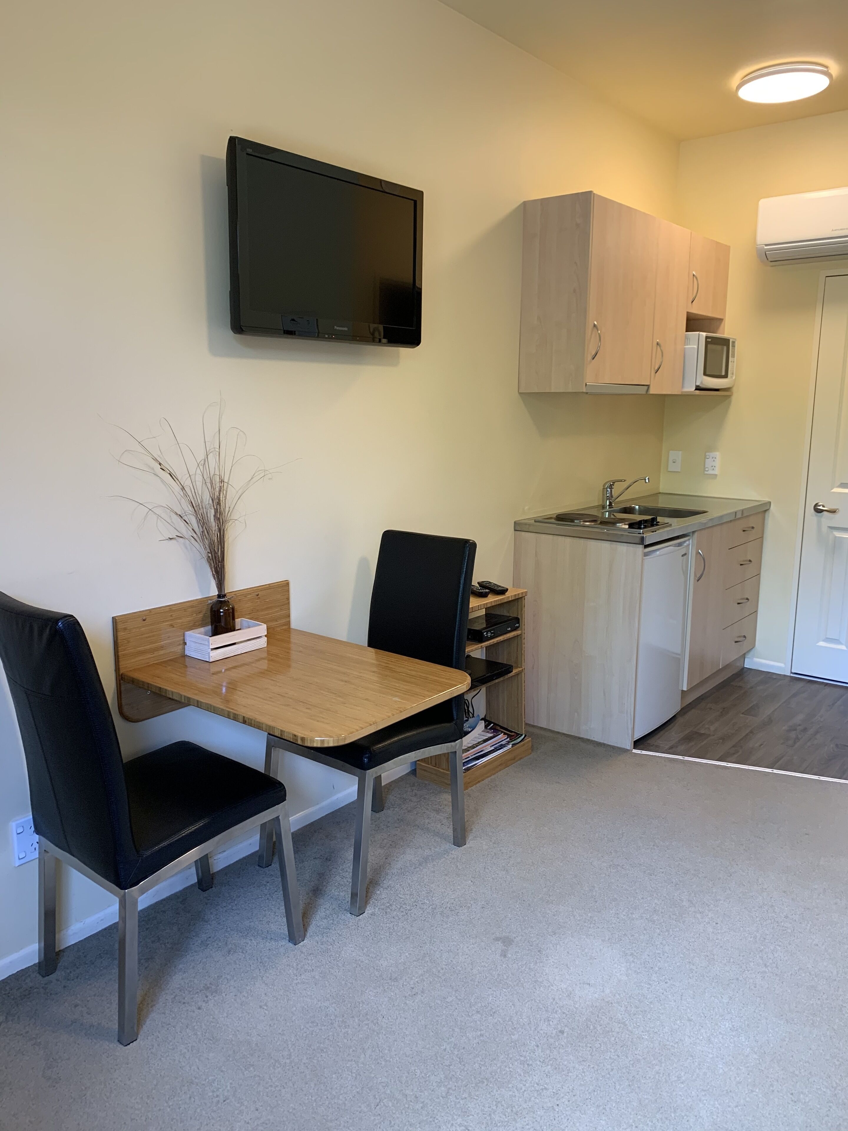 A compact kitchen with a microwave, stove, and sink. There is a dining table with chairs and a flat-screen TV mounted on the wall.