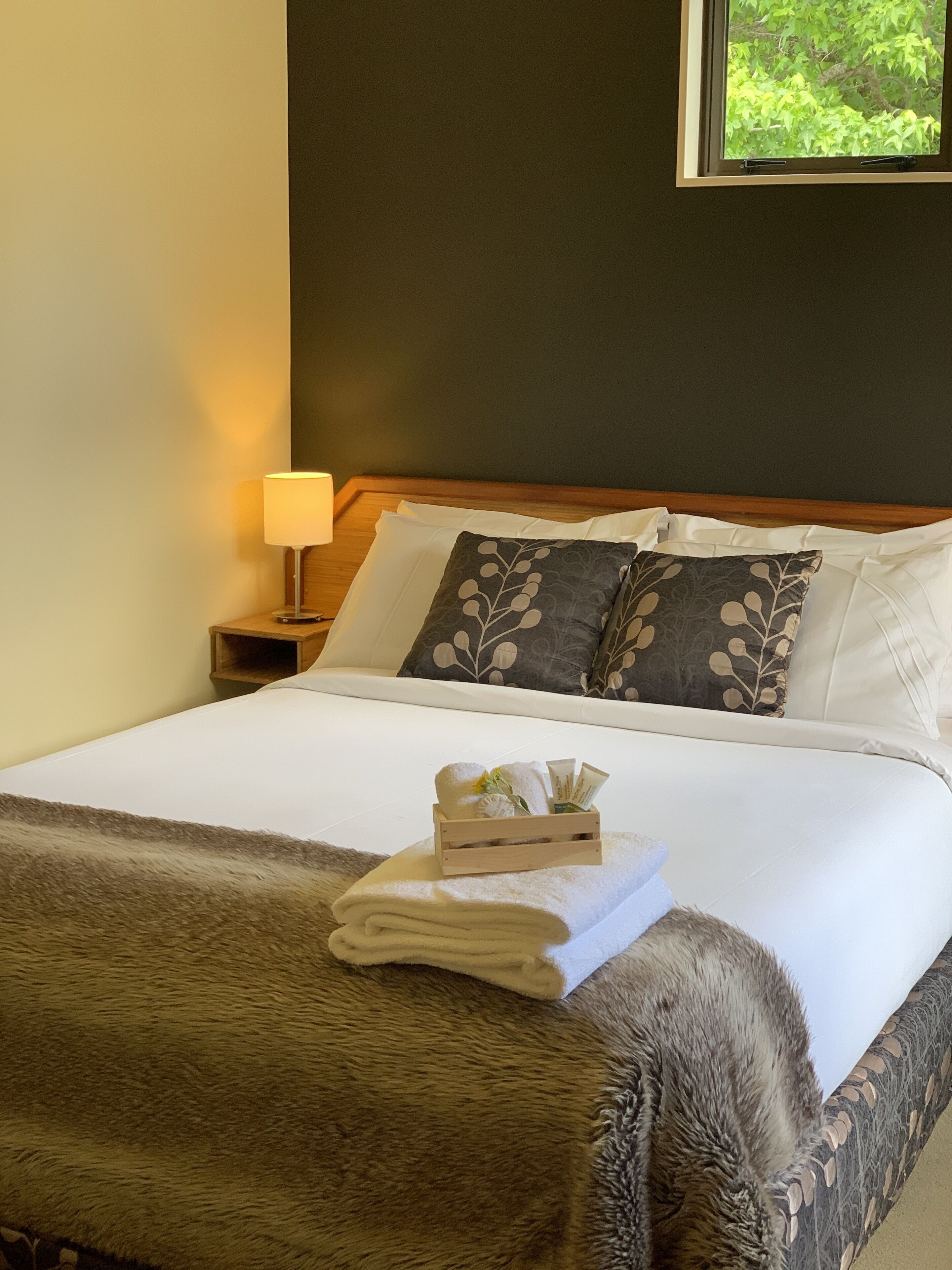 A neatly made bed with a white duvet, a brown throw blanket, and a wooden bedside table with a lamp.