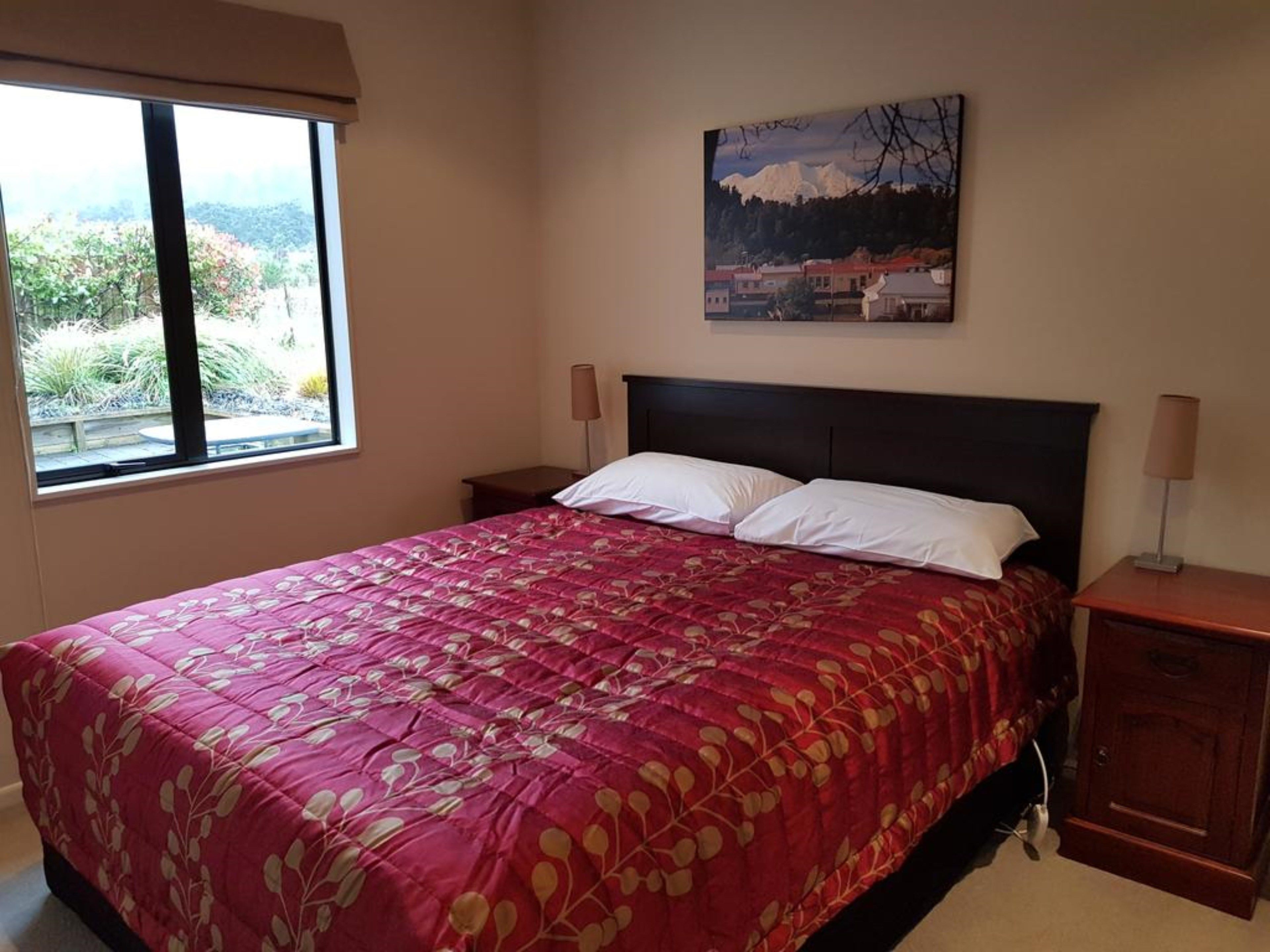 A bedroom with a large bed, two bedside tables, a window with a view of greenery, and a painting on the wall.