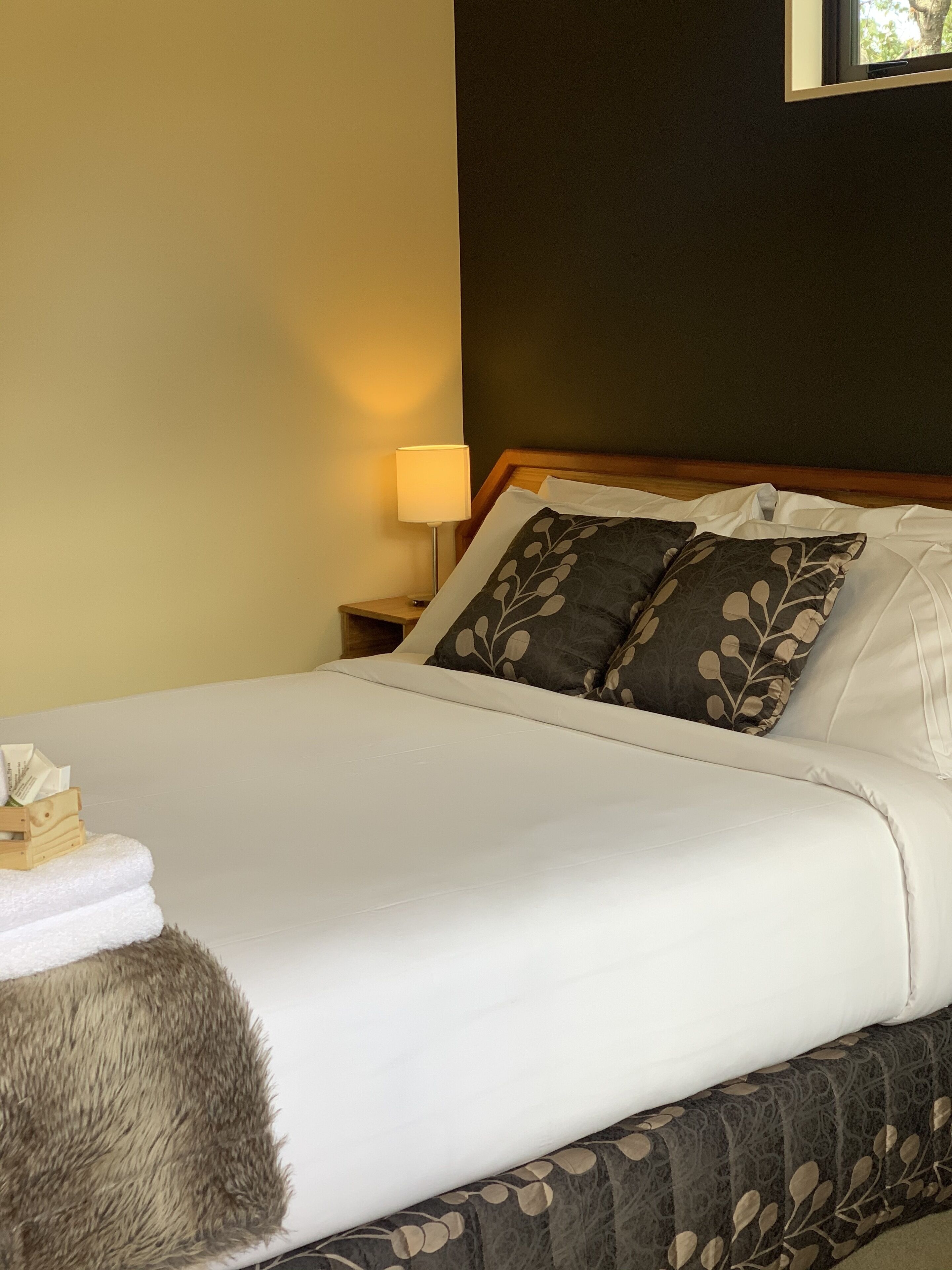 Una cama bien hecha con sábanas blancas y dos almohadones decorativos, una mesita de noche de madera con una lámpara, y una alfombra mullida al lado de la cama.