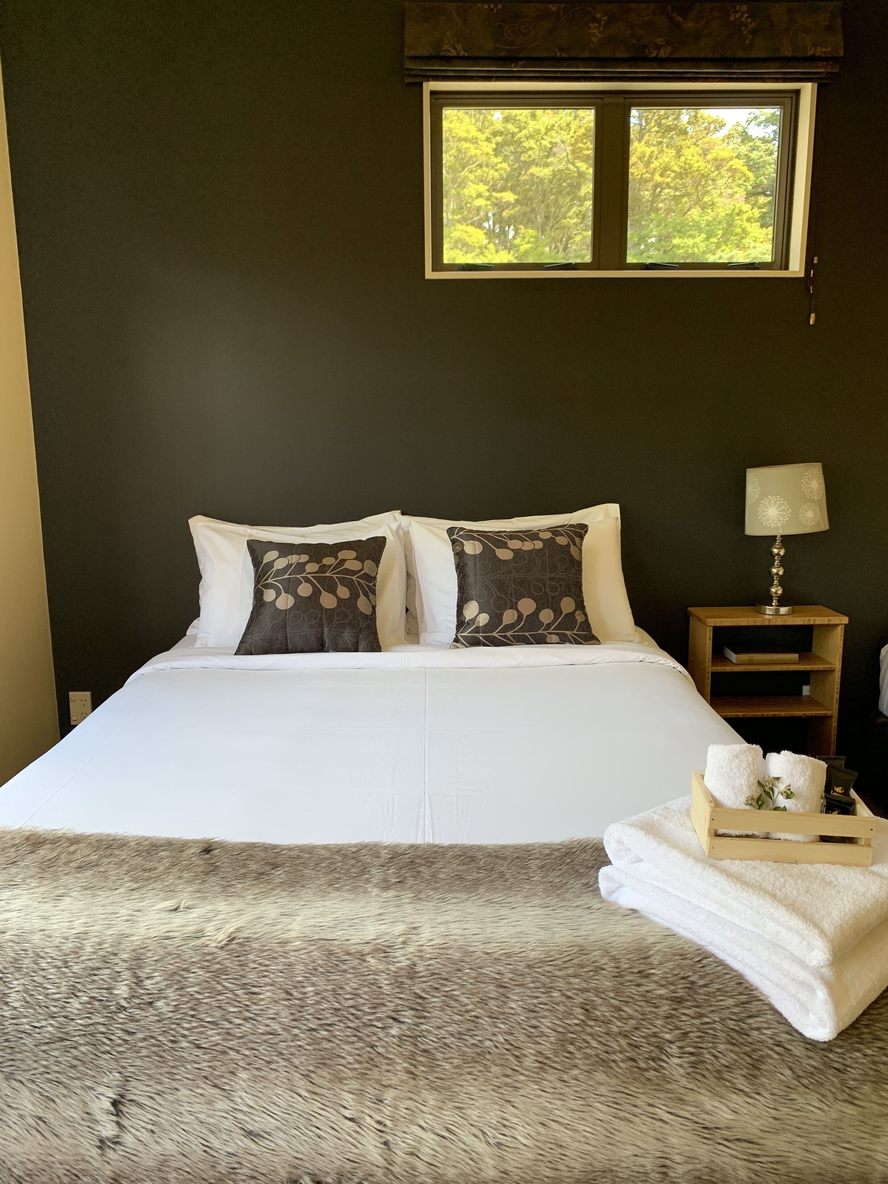 Una cama bien hecha con sábanas blancas y almohadones estampados, una lámpara de noche y una bandeja con toallas sobre una pared oscura con una ventana arriba.