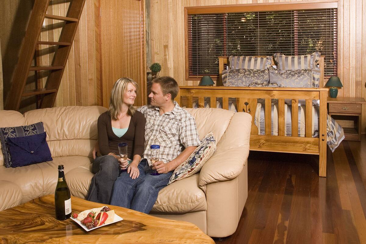 Un interior de madera con un sofá, una mesa de madera con una botella y aperitivos, y una ventana con persianas.