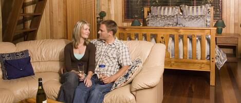 Un interior de madera con un sofá, una mesa de madera con una botella y aperitivos, y una ventana con persianas.