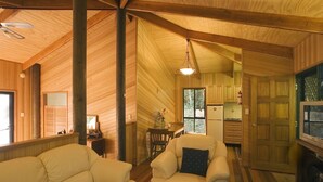L’intérieur chaleureux d’un chalet comprend un salon avec un canapé et un fauteuil, un coin repas avec une table et des chaises, une cuisine équipée de placards et d’un réfrigérateur, ainsi qu’un téléviseur posé sur un meuble.