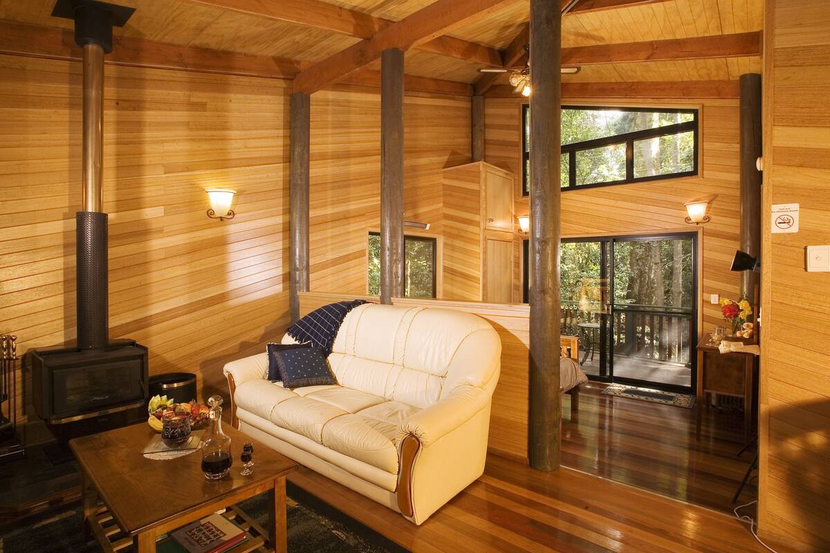 Interior acogedor de una cabaña con sofá, una mesa con bebidas, chimenea y ventanas amplias.