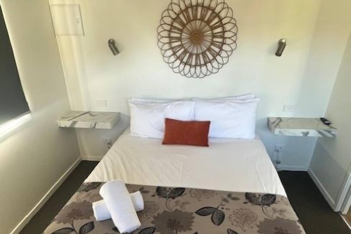 A neatly made bed with a floral bedspread, a decorative wall hanging, and a single red pillow.