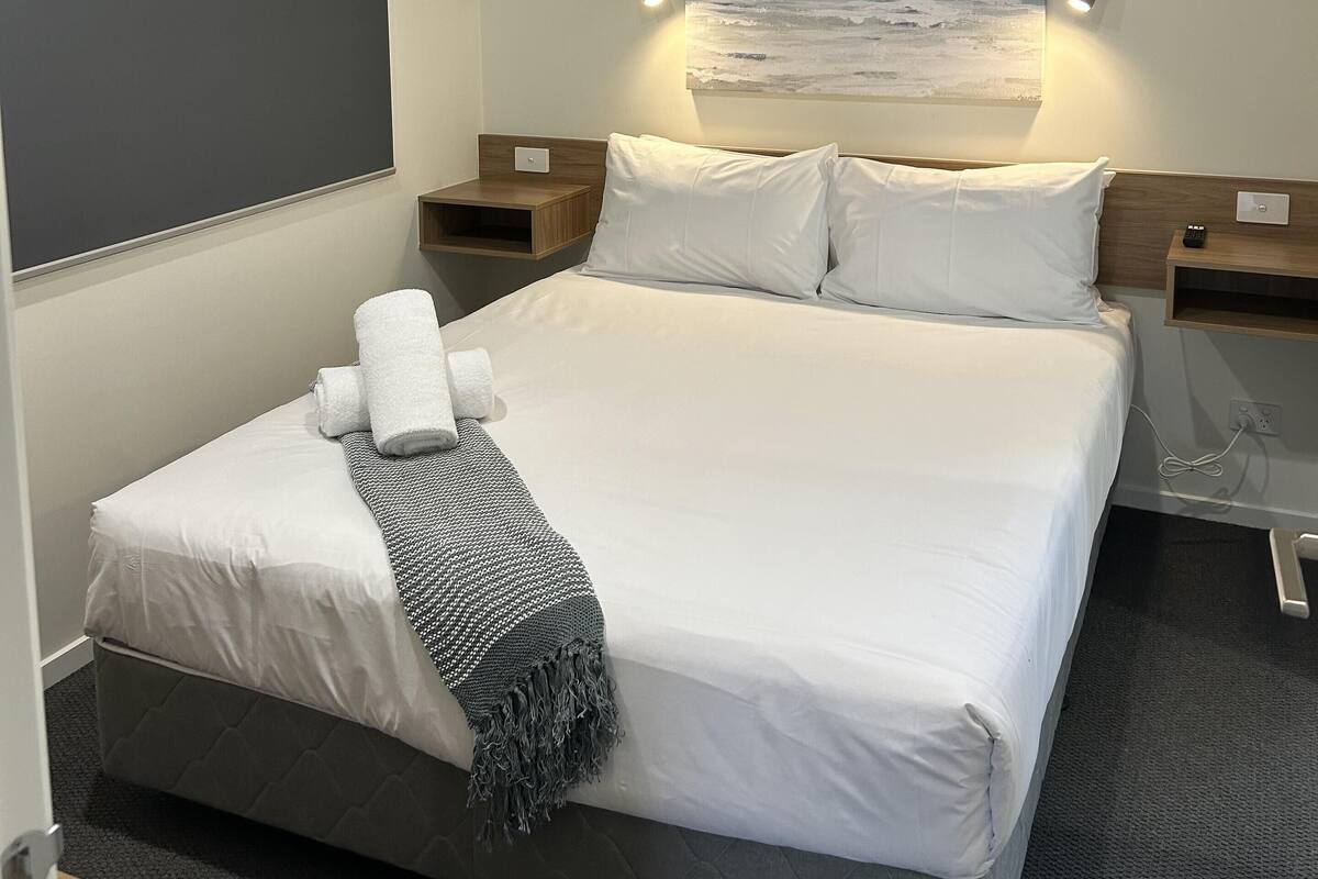 A neatly made bed with white linens and a gray blanket, a ceiling fan, a wall-mounted painting of the ocean, and bedside tables with lamps.