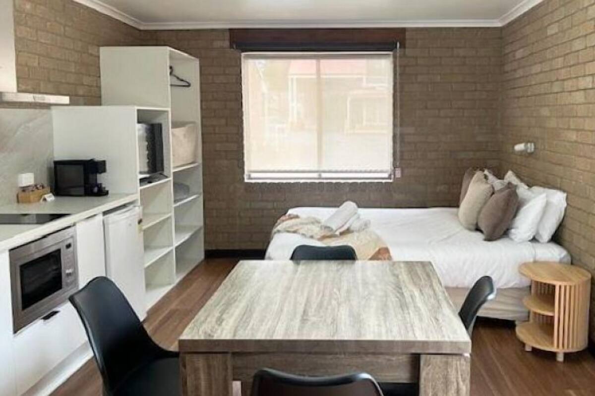 A compact room with a kitchenette, a bed with pillows, a small round table, and a window with blinds.
