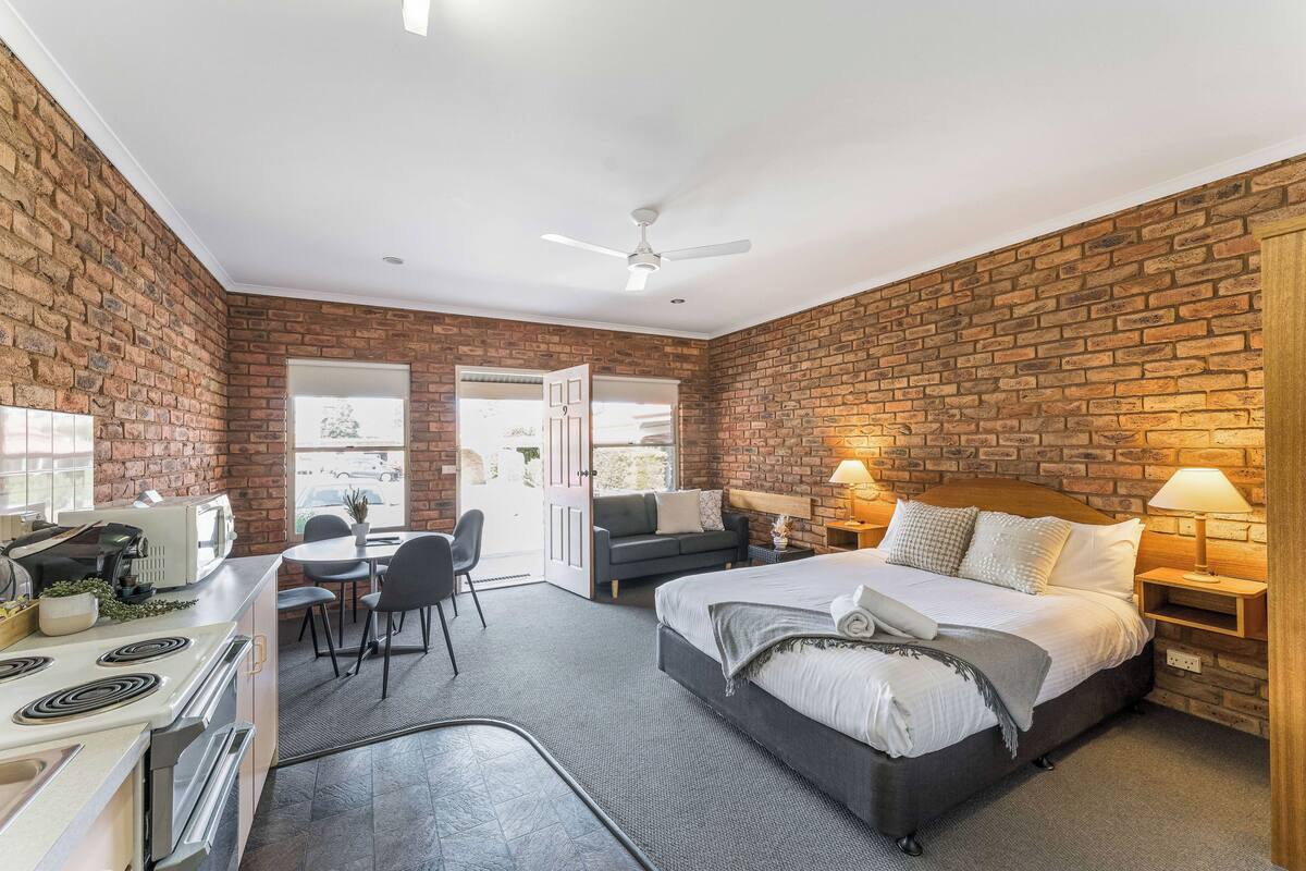 A compact hotel room with a brick wall, a bed, a desk with a chair, a small kitchenette, and a ceiling fan.