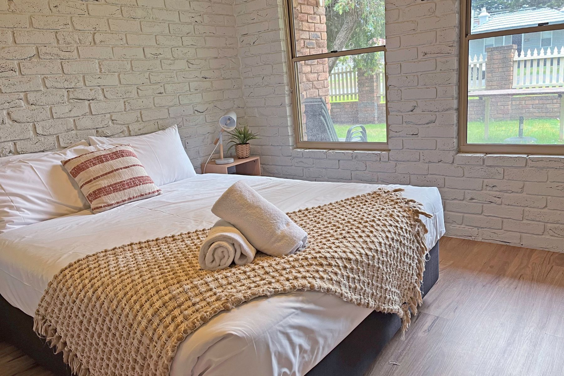 Ein Schlafzimmer mit einer Ziegelwand, einem großen Bett mit weißer Bettwäsche, einer gewobenen Tagesdecke und Blick auf einen Garten durch ein Fenster.
