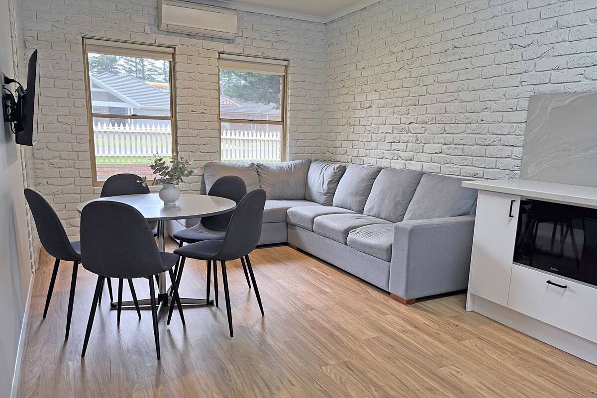 A compact living space with a grey sofa, a round dining table with four chairs, and a white cabinet with a built-in oven.