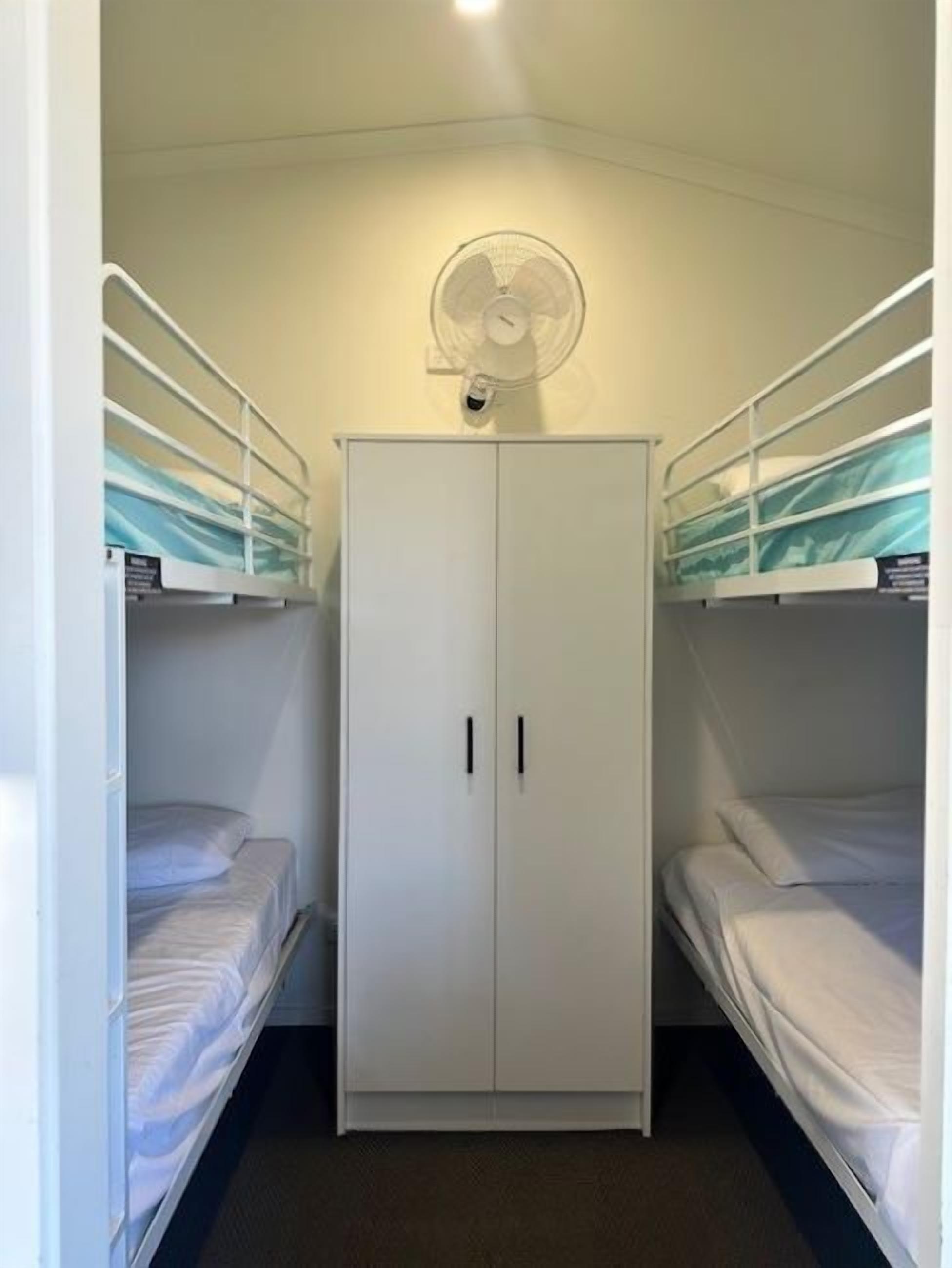 A small, compact room with two bunk beds, a white wardrobe, and a ceiling fan.
