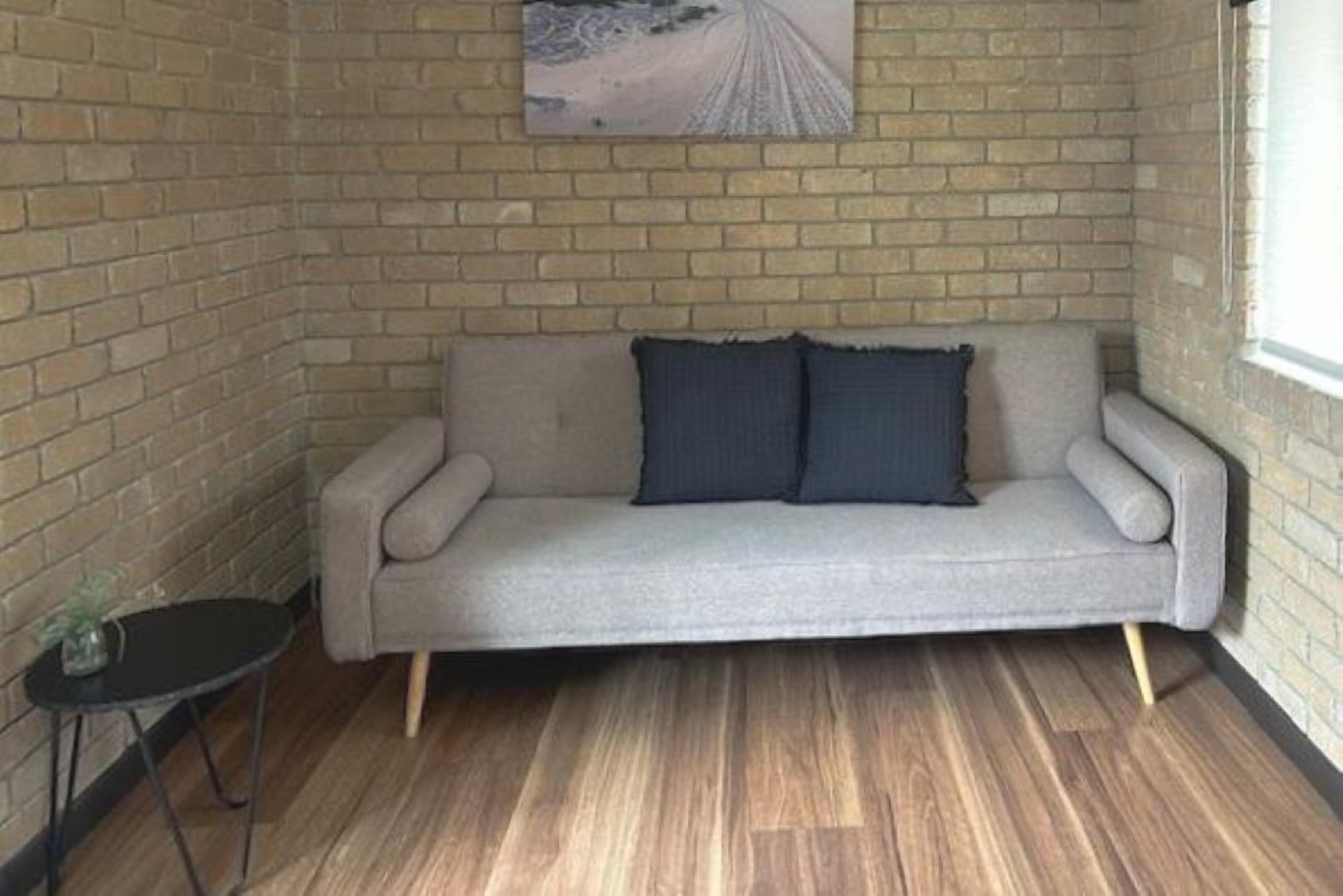A corner room with a brick wall, a grey sofa with two cushions, a small black table, and a window.