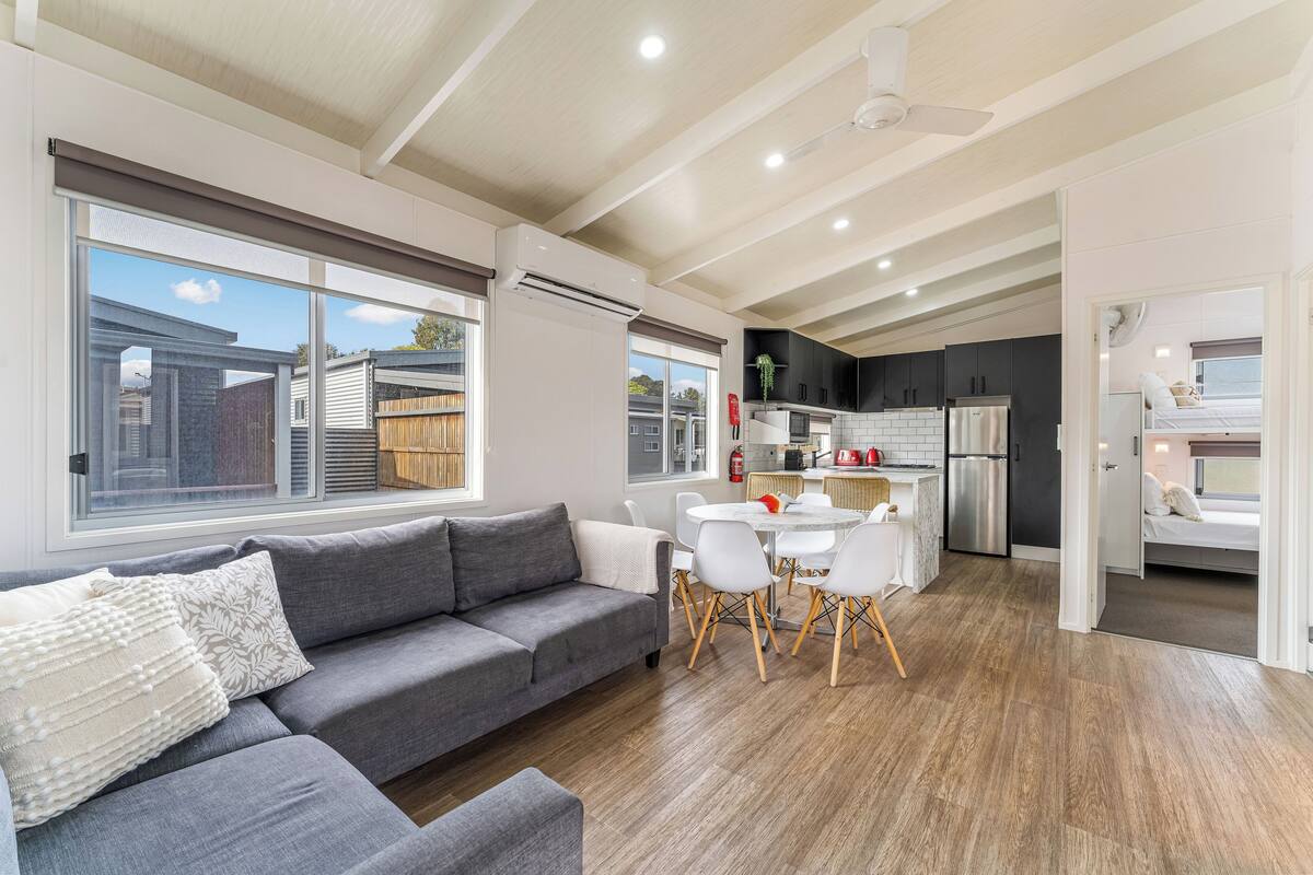 A modern living space with a grey sofa, dining table, and chairs, a kitchen with dark cabinets, and a large window.