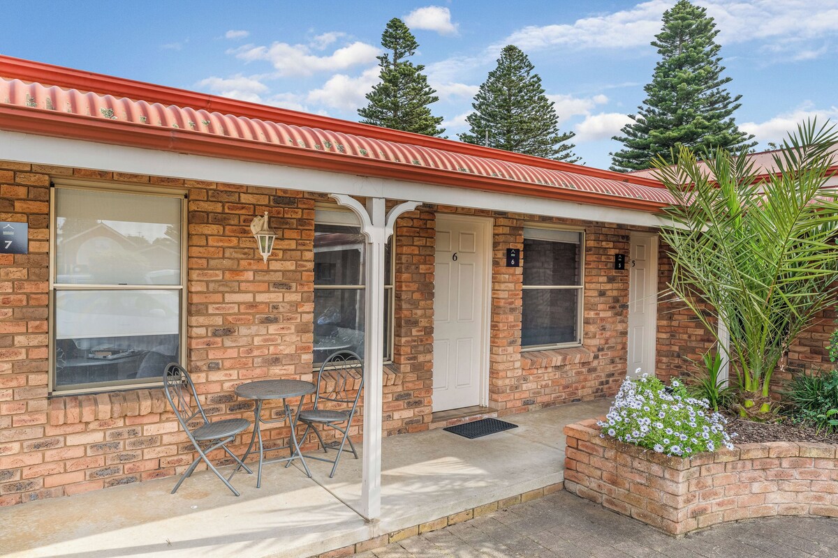 A brick building with a red roof, numbered doors, and a small patio with chairs.