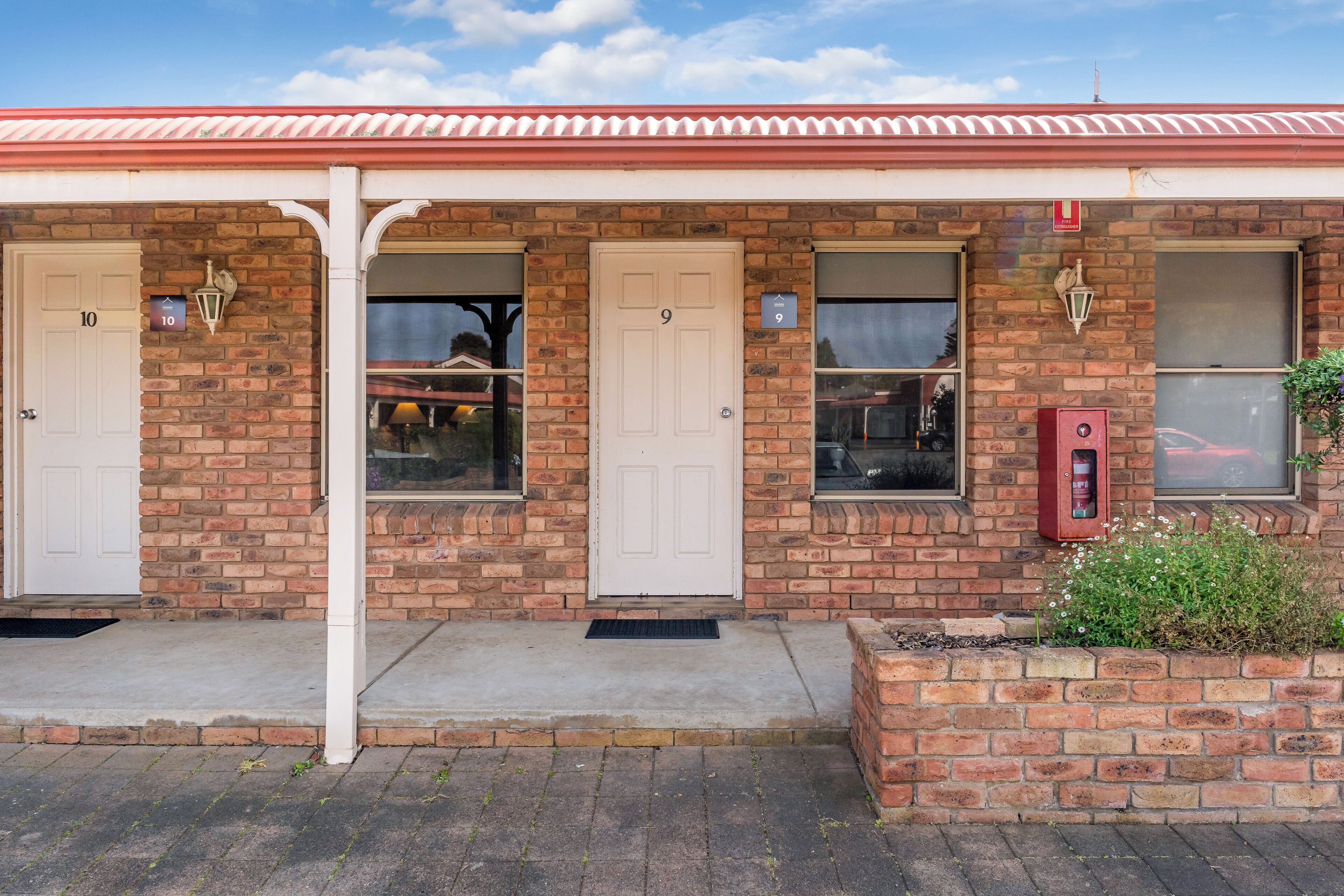 A two-story brick building with numbered doors, a red mailbox, and a small garden area.