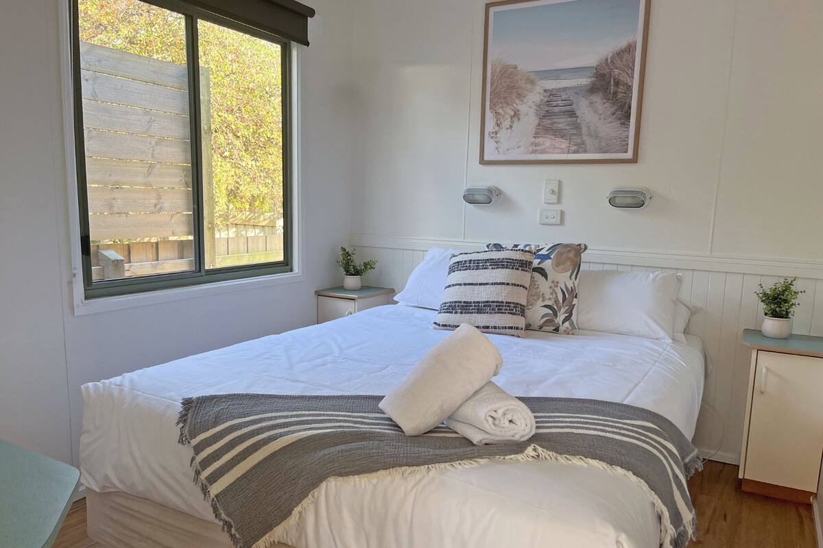 A neatly made bed with a striped blanket, a rolled towel, and decorative pillows. There is a window with a view of trees and a framed picture on the wall.
