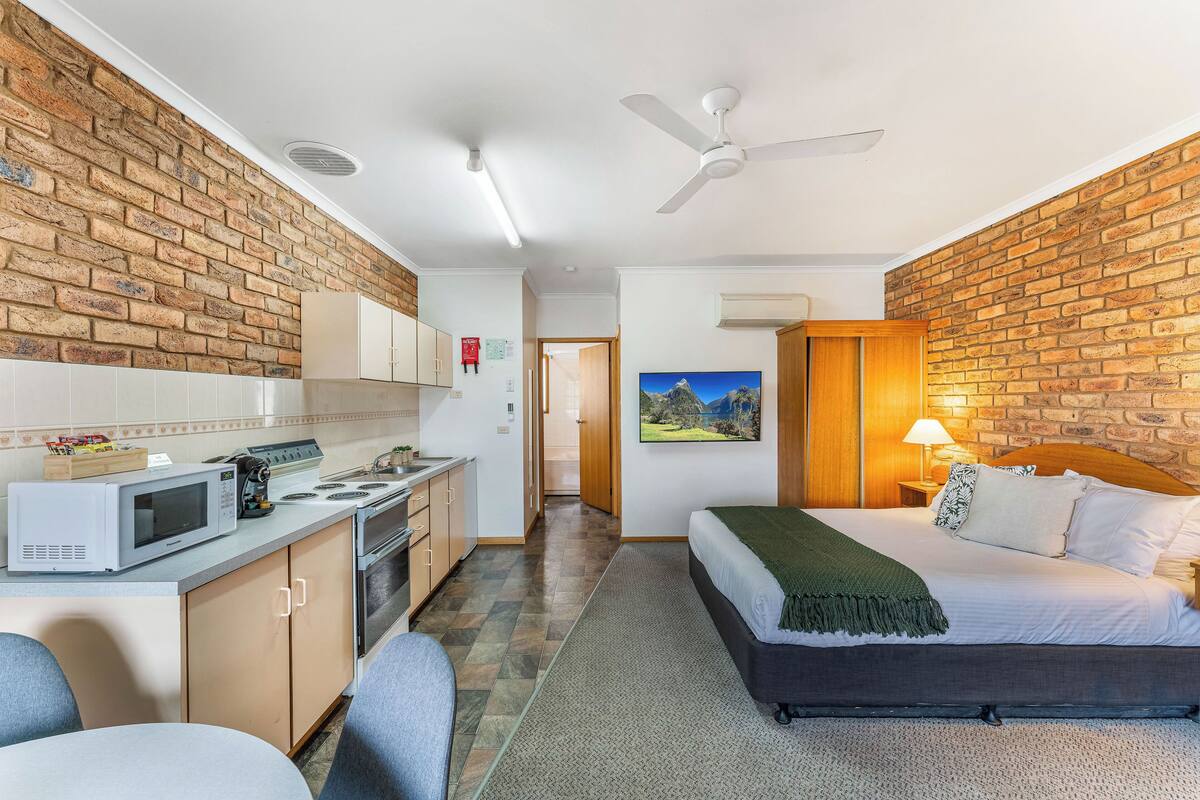 A compact hotel room with a kitchenette, a bed with a green throw, and a brick wall.