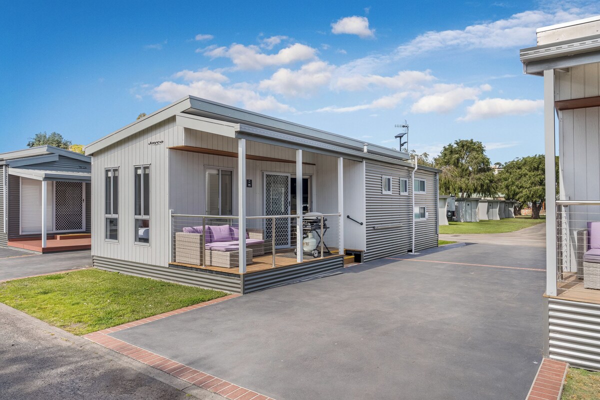 A modern mobile home with a covered porch, a small patio, and a well-maintained lawn.