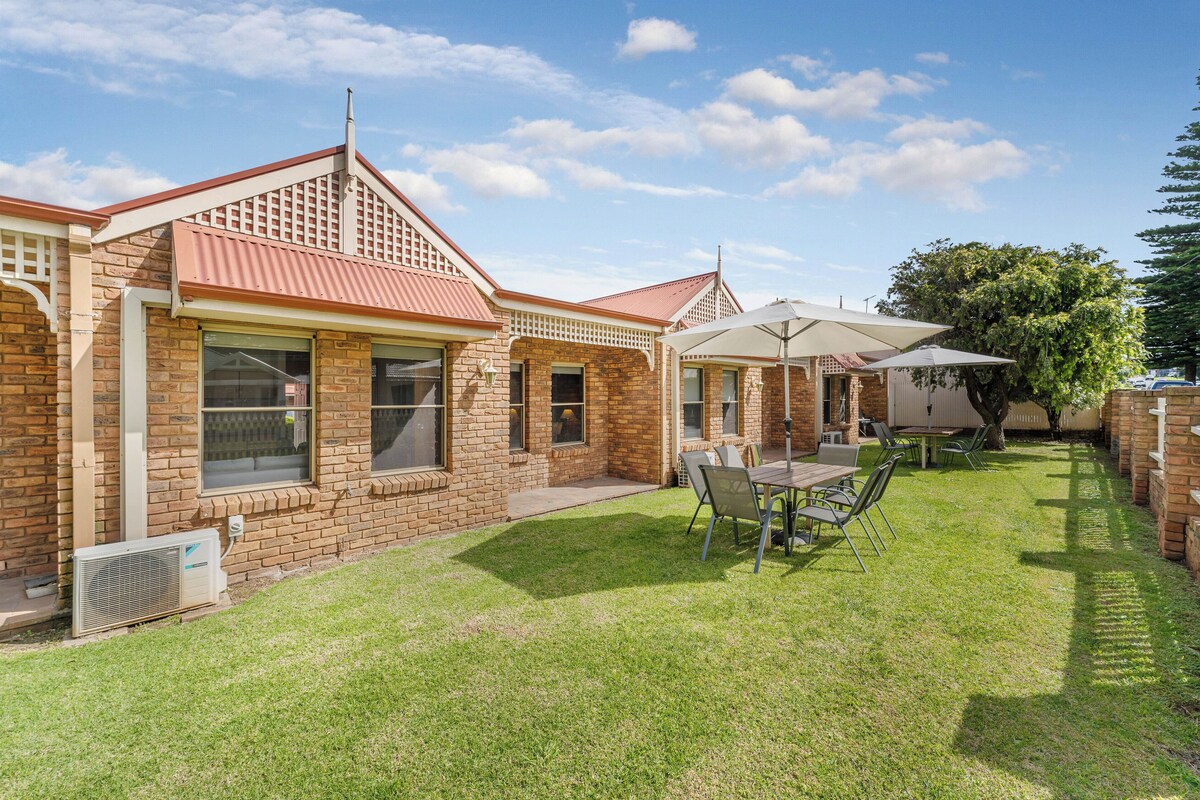 A brick house with a red roof, a patio with a table and chairs, and a lawn.