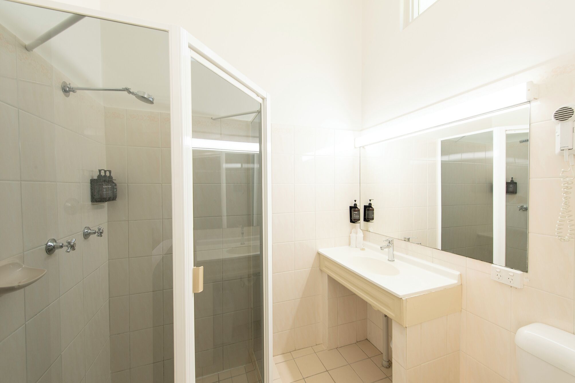 Une salle de bain avec une douche à parois de verre, un lavabo blanc avec un miroir et deux distributeurs de savon.