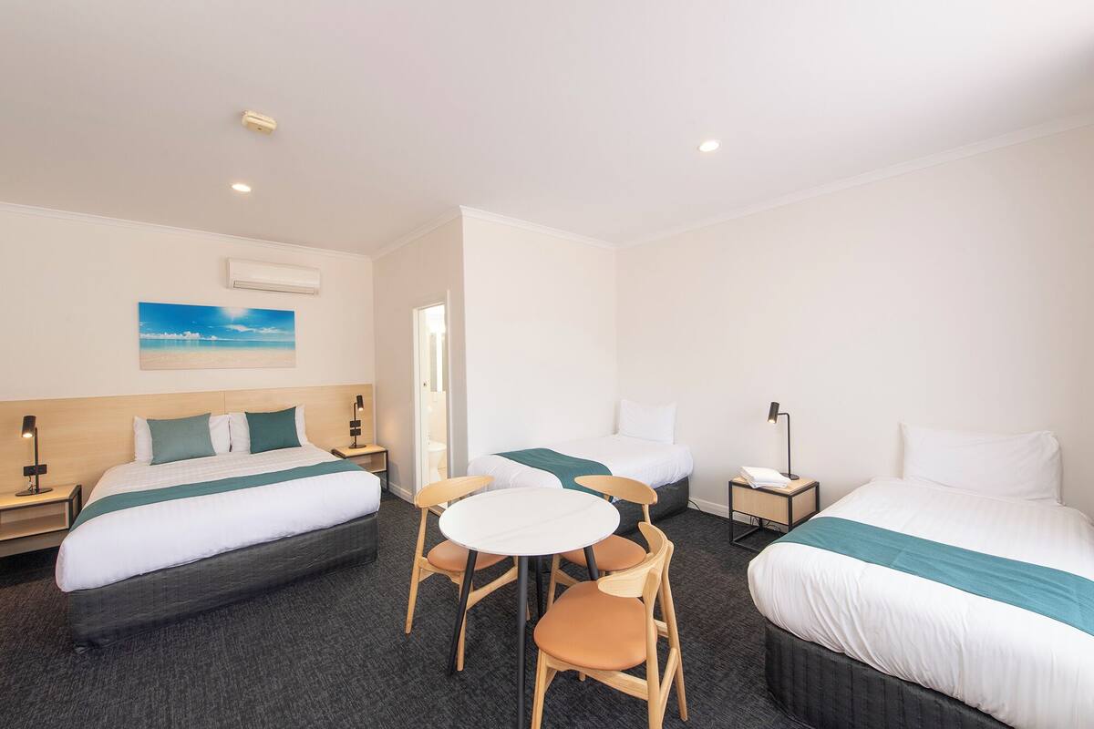 A hotel room with two beds, a small table, and chairs. There is a painting on the wall and a door leading to another room.