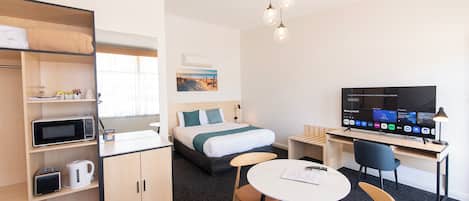 A hotel room with a bed, a desk with a TV, a dining table with chairs, a microwave, a kettle, and a shelf with a kettle and a toaster.