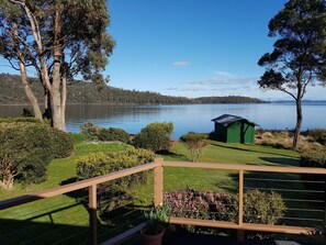 A lakeside view with a wooden deck, green shed, and various shrubs. - Abs by the Bay (Taranna)