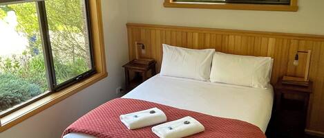 Un lit bien fait, recouvert d’une couverture rouge, avec deux oreillers et deux serviettes. Il y a une fenêtre sur le côté gauche de la pièce, une tête de lit en bois et une petite table de chevet avec une lampe.