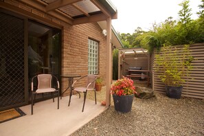 A patio area with two wicker chairs, a potted plant with red flowers, and a brick building with a sliding glass door.