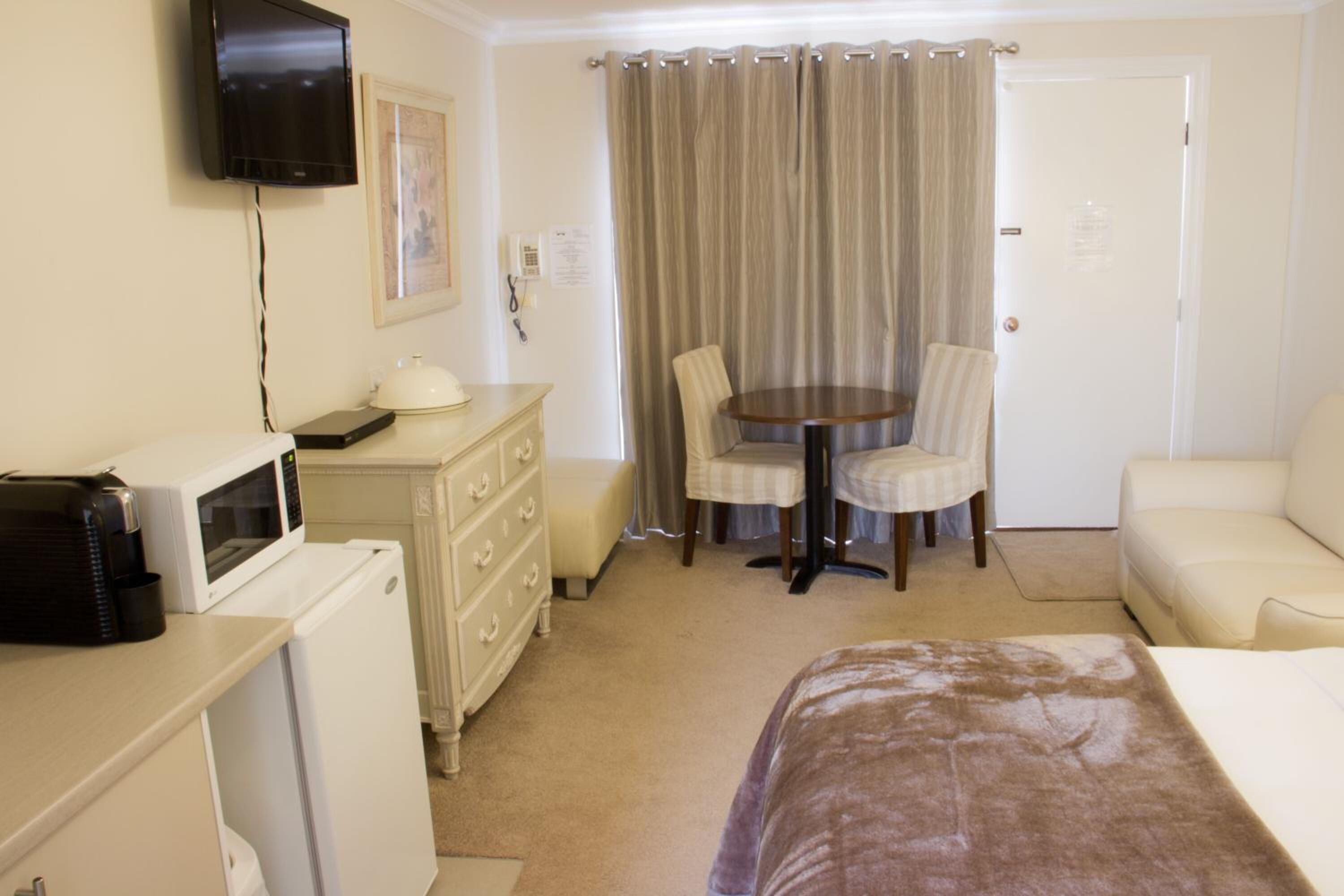 A hotel room with a bed, a desk with a microwave, a dresser, a television, a dining table with chairs, and a door.