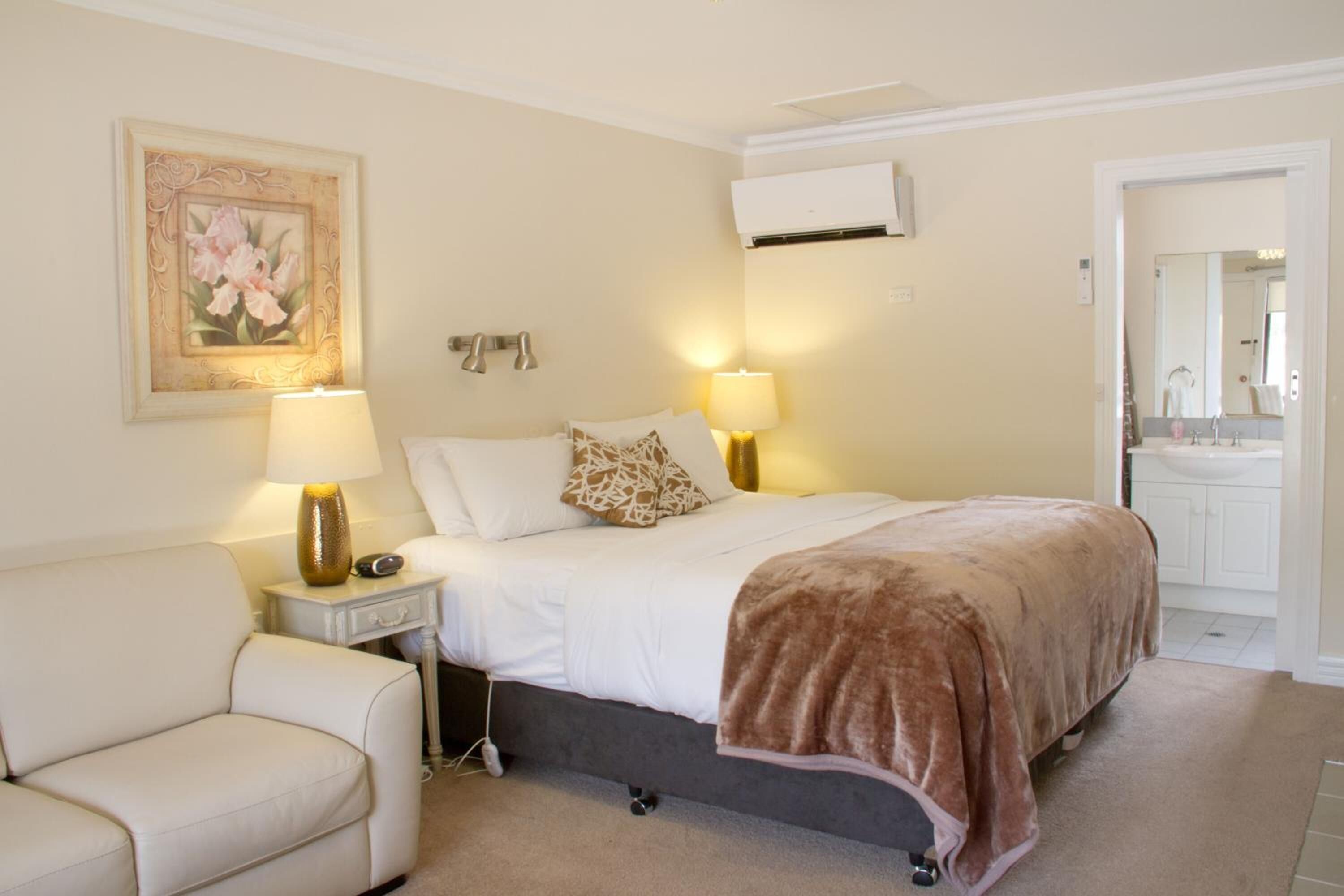 Habitación de hotel con cama, lámparas de noche, mesita de noche, una silla, un cuadro, equipo de aire acondicionado y baño visible a través de una puerta abierta.