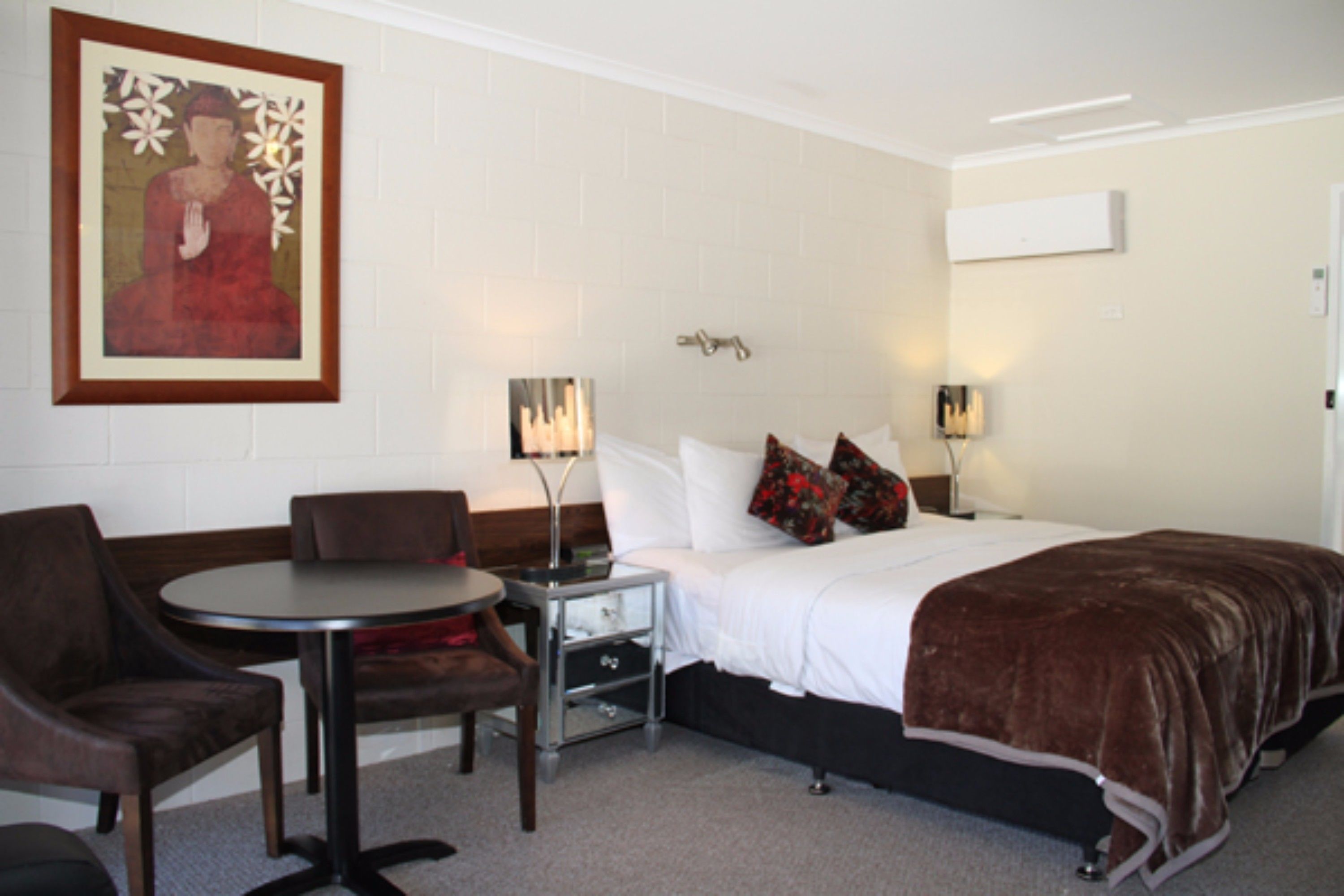 A hotel room with a bed, a small table with chairs, a nightstand, and a wall-mounted air conditioner.