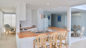 A modern kitchen with a central island, white cabinetry, and a tiled backsplash. There are wicker chairs around the island and a dining table with chairs in the adjacent room. - Ultimate Holiday Apartments (Lakes Entrance)