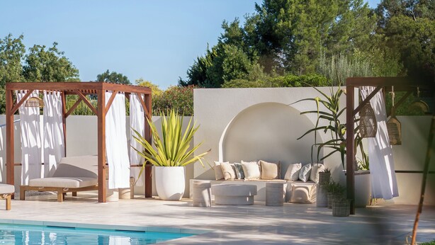 Un área junto a la piscina con una pérgola de madera, sillones de descanso, un sofá y una planta en maceta.