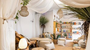 Una habitación acogedora y bien iluminada, con piso de madera, un sofá cómodo, un rincón de lectura y una estantería llena de libros y objetos decorativos.