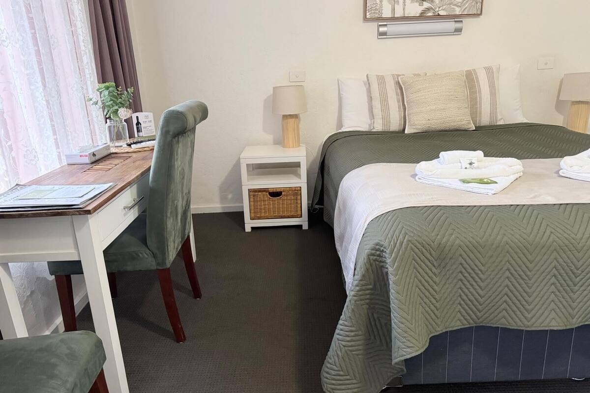 Habitación de hotel con cama, escritorio, silla, mesita de noche, lámpara y una obra de arte colgada en la pared.