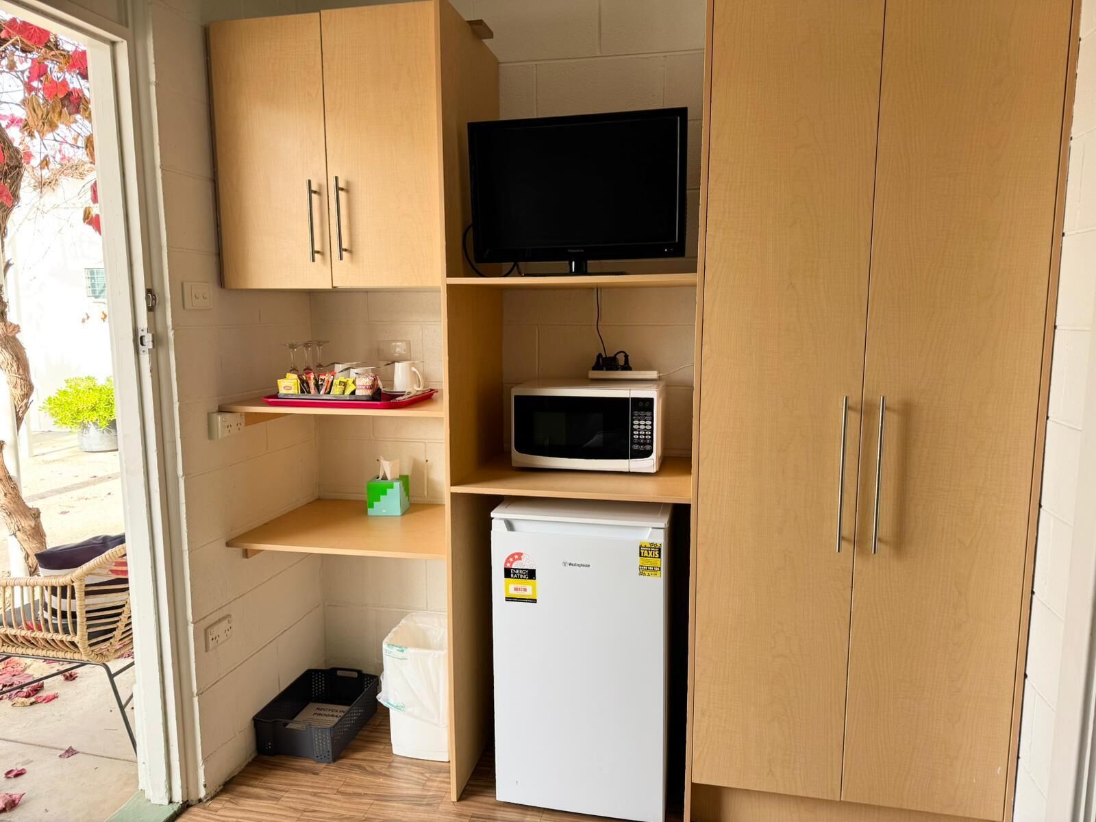 A compact kitchenette with wooden cabinets, a microwave, a small refrigerator, and a television on a shelf.