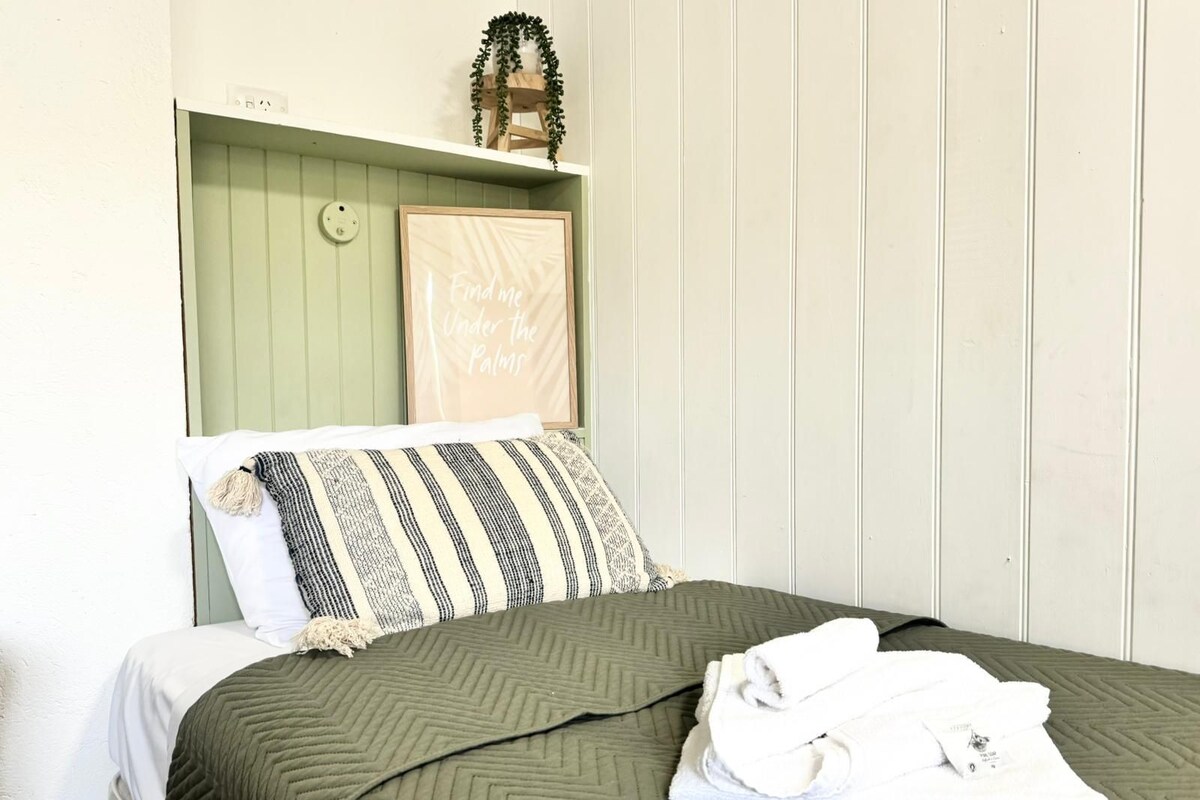 A bed with a green quilt, a striped pillow, and a folded towel on a white mattress.