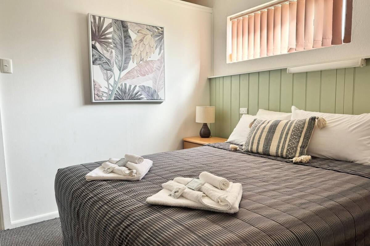 Una cama bien hecha con colcha y almohadas a rayas, una mesita de noche con lámpara y un cuadro con motivos tropicales en la pared.