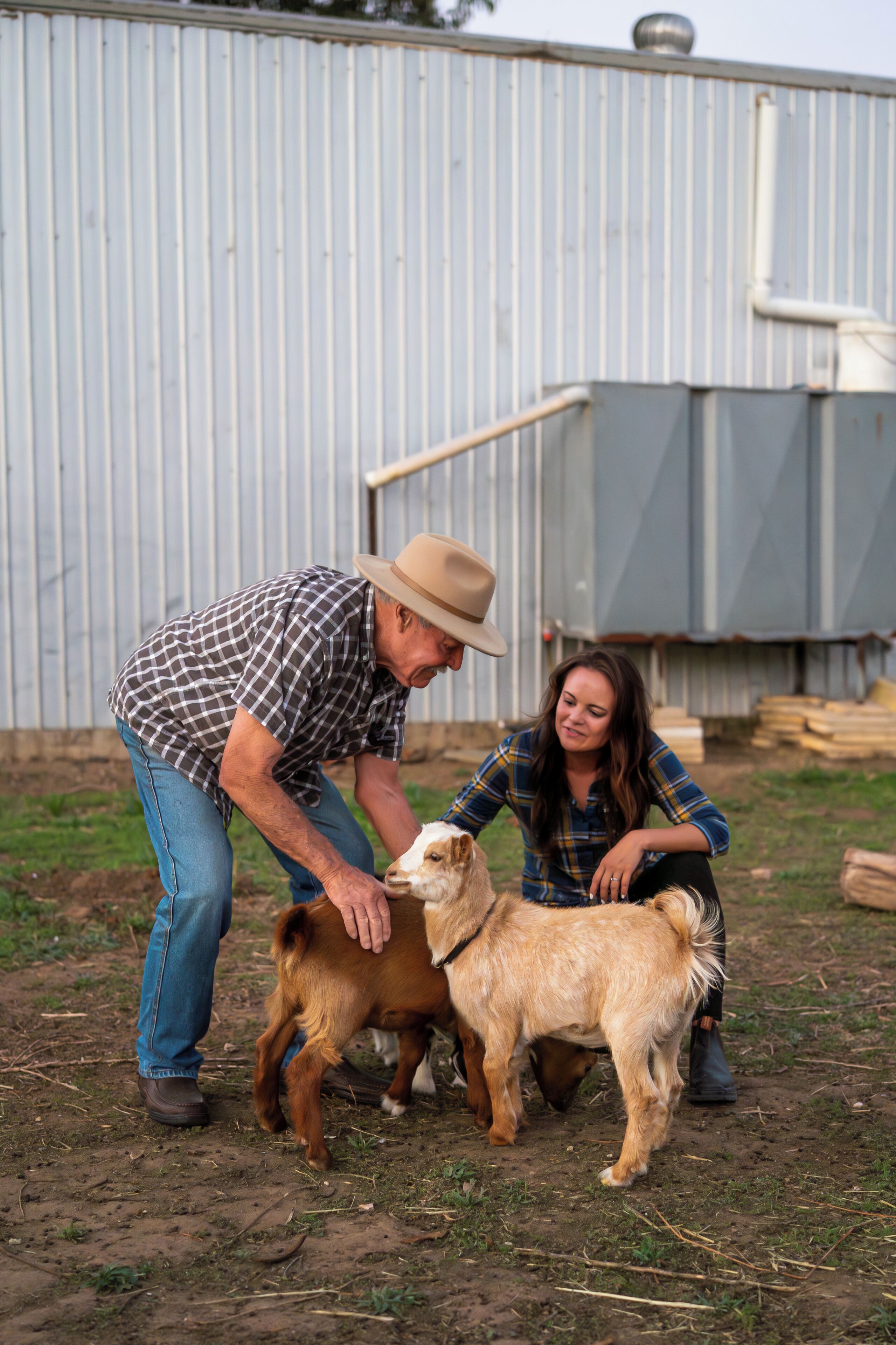 A man and a woman interacting with a small brown dog in an outdoor setting with a corrugated metal wall in the background.
