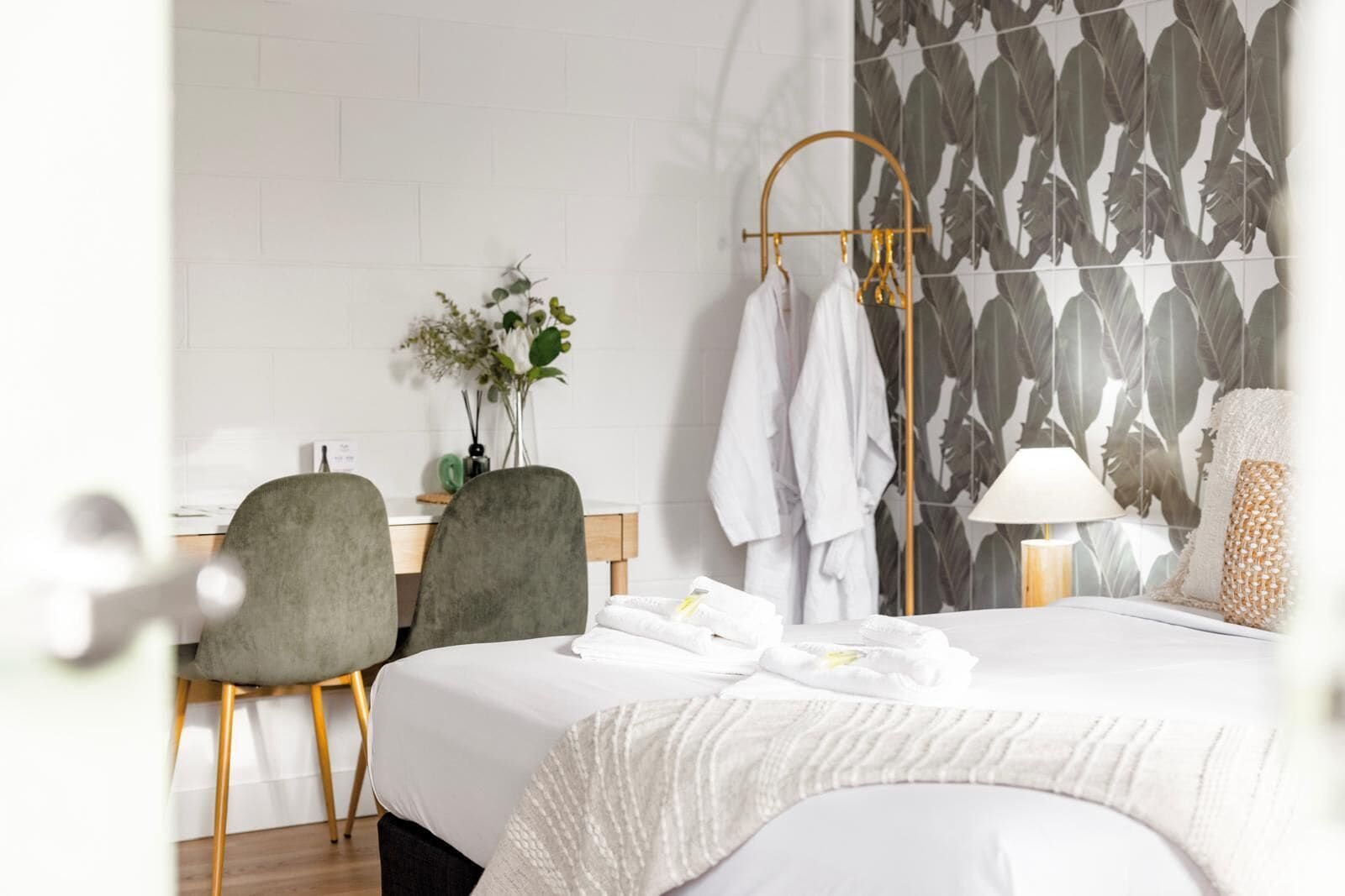 Una habitación de hotel con una cama, dos sillas, un escritorio, una planta, un perchero para toallas y una lámpara.
