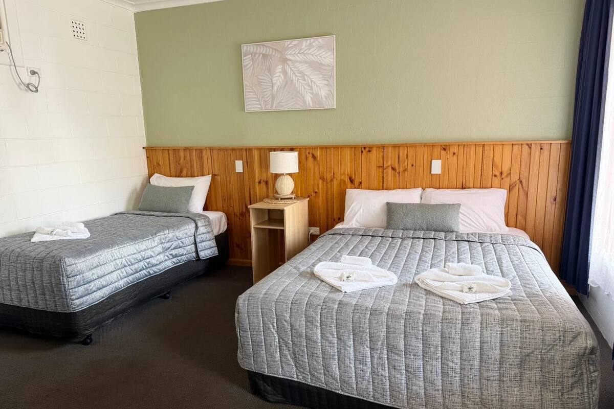 Habitación de hotel con dos camas, cabecera de madera, mesita de noche pequeña con lámpara y una obra de arte colgada en la pared.