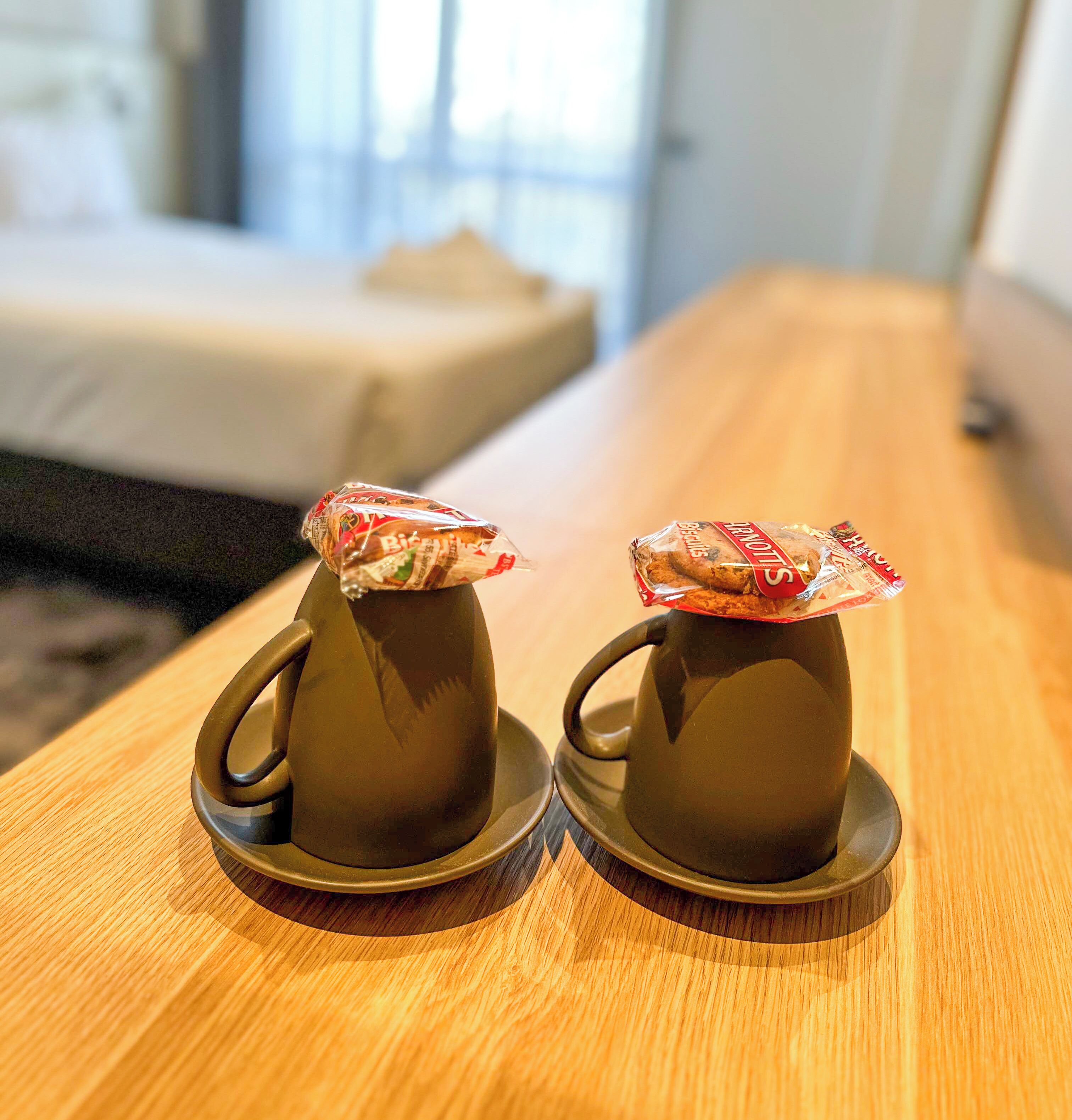 Two dark-colored cups with lids on a wooden table in a room with a bed in the background.
