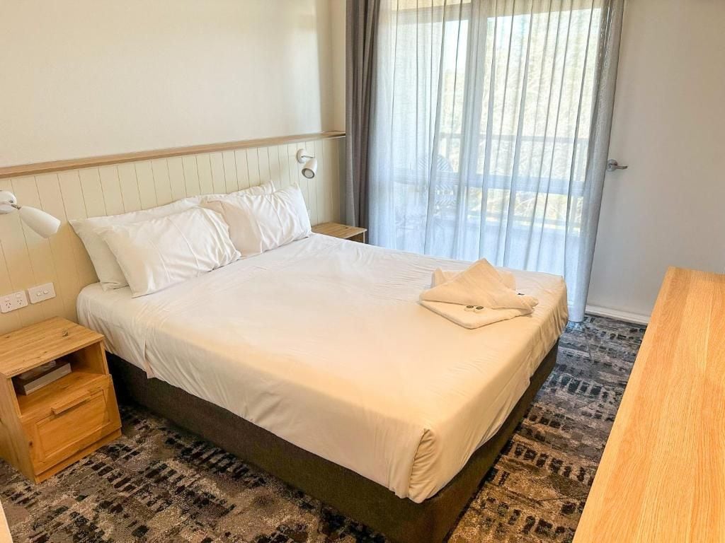 A hotel room with a bed, a nightstand, a wooden table, and a window with curtains.