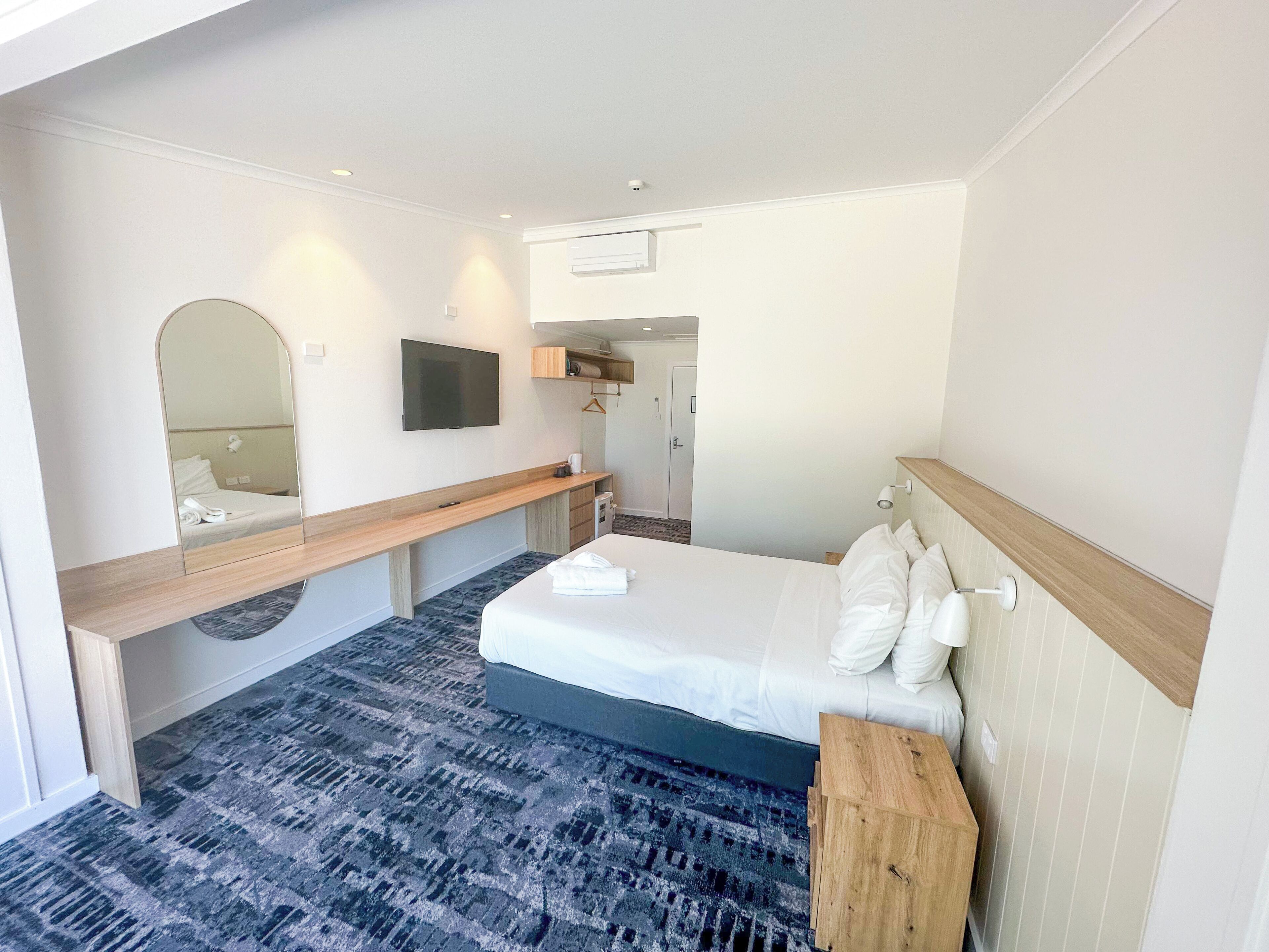 Une chambre d’hôtel avec un lit, une table de chevet, un miroir, une télévision et une salle de bain visible à travers une porte ouverte.